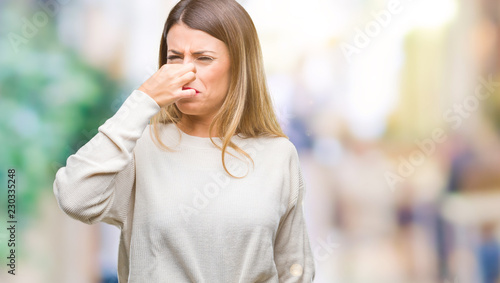 Young beautiful woman casual white sweater over isolated background smelling something stinky and disgusting, intolerable smell, holding breath with fingers on nose. Bad smells concept.