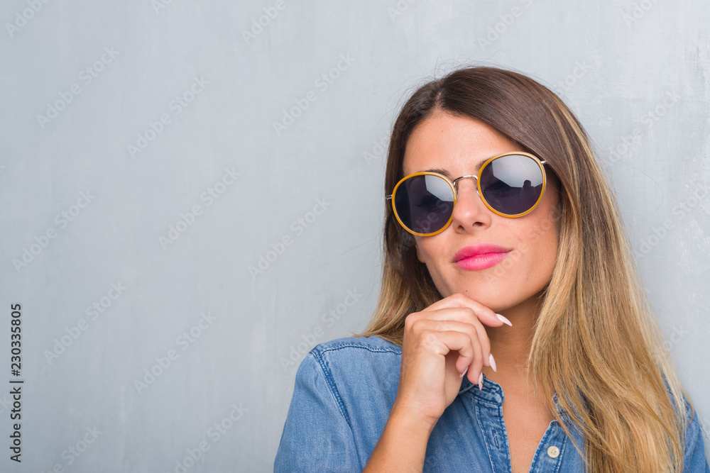 Young adult woman over grunge grey wall wearing retro sunglasses with hand on chin thinking about question, pensive expression. Smiling with thoughtful face. Doubt concept. Young adult woman over grunge grey wall wearing retro sunglasses with hand on chin thinking about question, pensive expression. Smiling with thoughtful face. Doubt concept.