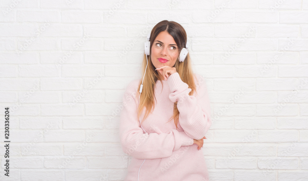 Young adult woman over white brick wall wearing headphones listening to music and dancing serious face thinking about question, very confused idea Young adult woman over white brick wall wearing headphones listening to music and dancing serious face thinking about question, very confused idea