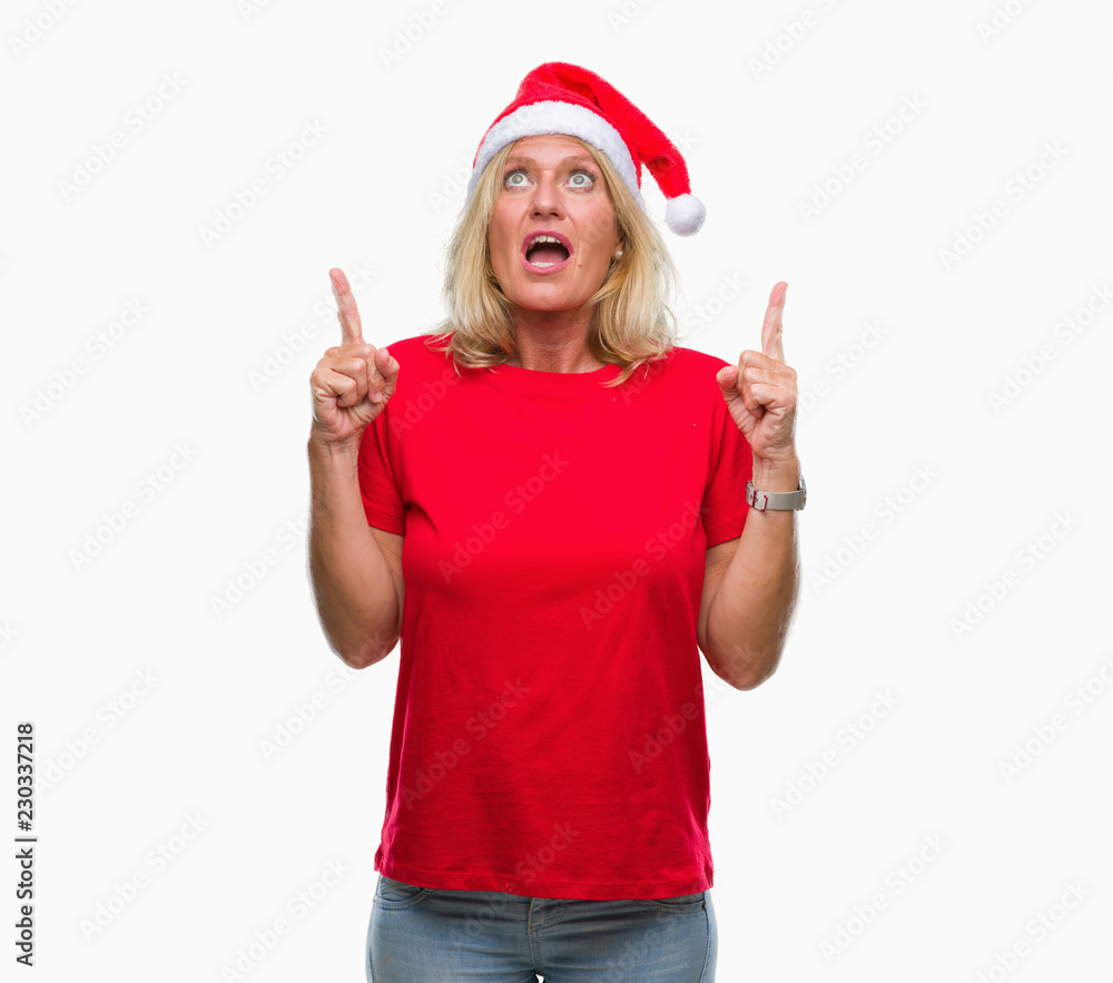 Middle age blonde woman wearing christmas hat over isolated background amazed and surprised looking up and pointing with fingers and raised arms. Middle age blonde woman wearing christmas hat over isolated background amazed and surprised looking up and pointing with fingers and raised arms.