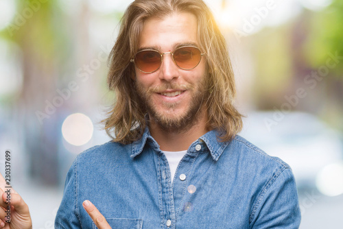 Young handsome man with long hair wearing sunglasses over isolated background smiling and looking at the camera pointing with two hands and fingers to the side.