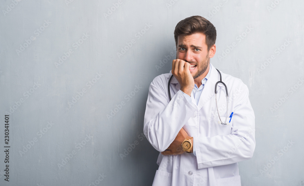 Handsome young doctor man over grey grunge wall looking stressed and nervous with hands on mouth biting nails. Anxiety problem. Handsome young doctor man over grey grunge wall looking stressed and nervous with hands on mouth biting nails. Anxiety problem.