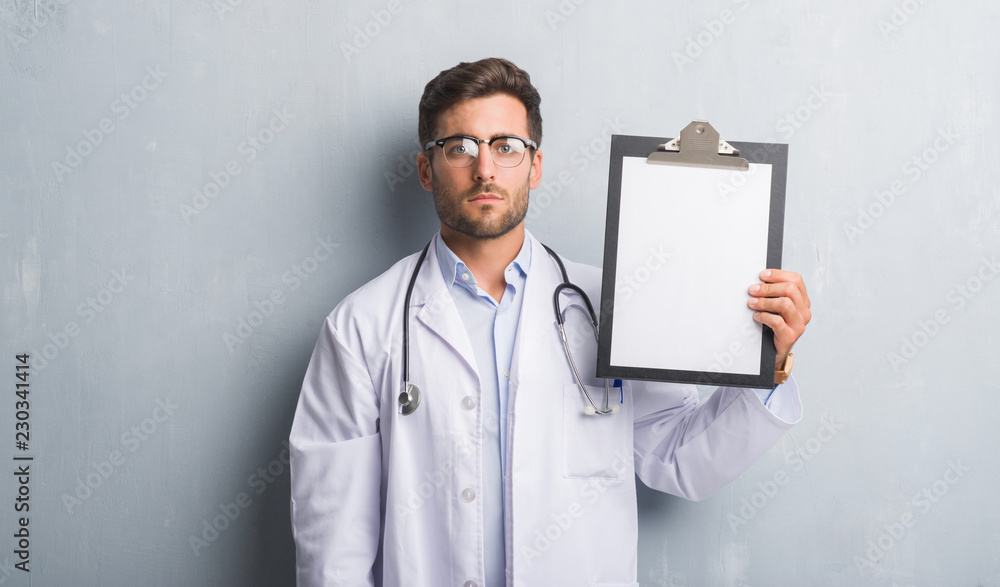 Handsome young doctor man over grey grunge wall holding clipboard with a confident expression on smart face thinking serious Handsome young doctor man over grey grunge wall holding clipboard with a confident expression on smart face thinking serious