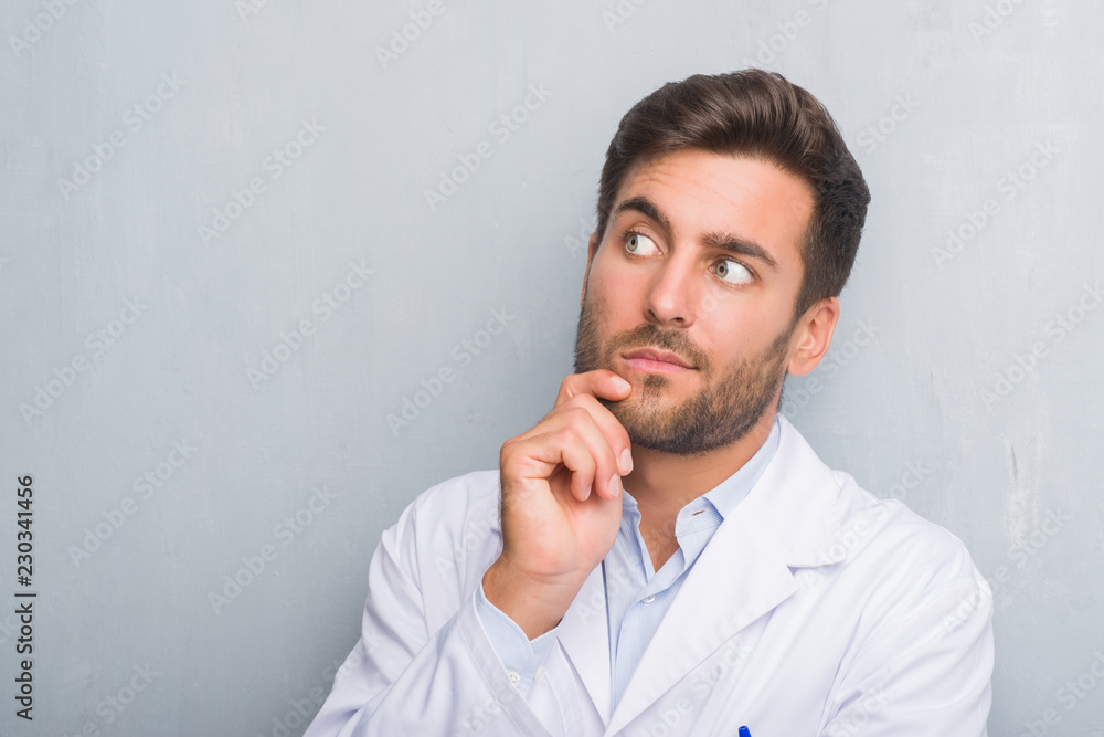 Handsome young professional man over grey grunge wall wearing white coat with hand on chin thinking about question, pensive expression. Smiling with thoughtful face. Doubt concept. Handsome young professional man over grey grunge wall wearing white coat with hand on chin thinking about question, pensive expression. Smiling with thoughtful face. Doubt concept.