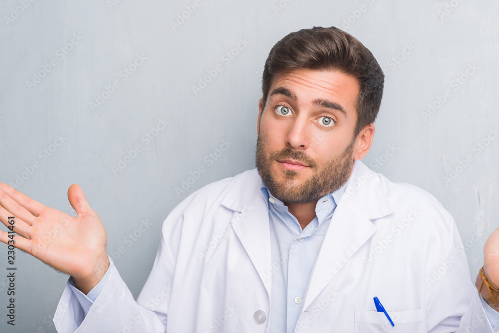 Handsome young professional man over grey grunge wall wearing white coat clueless and confused expression with arms and hands raised. Doubt concept. Handsome young professional man over grey grunge wall wearing white coat clueless and confused expression with arms and hands raised. Doubt concept.