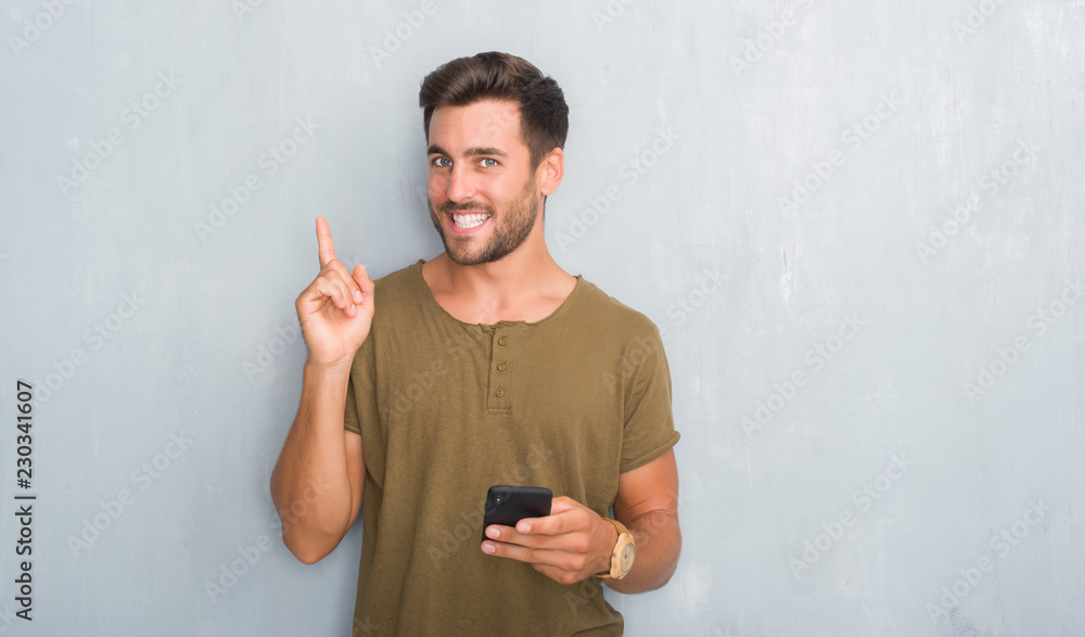 Handsome young man over grey grunge wall texting a message using smartphone very happy pointing with hand and finger to the side Handsome young man over grey grunge wall texting a message using smartphone very happy pointing with hand and finger to the side