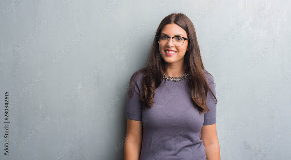 Young brunette woman over grunge grey wall wearing glasses with a happy face standing and smiling with a confident smile showing teeth Young brunette woman over grunge grey wall wearing glasses with a happy face standing and smiling with a confident smile showing teeth