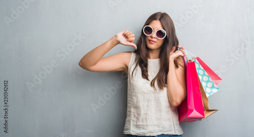 Young brunette woman over grunge grey wall holding colorful shopping bags with angry face, negative sign showing dislike with thumbs down, rejection concept