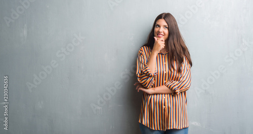 Young brunette woman over grunge grey wall looking confident at the camera with smile with crossed arms and hand raised on chin. Thinking positive. © Krakenimages.com
