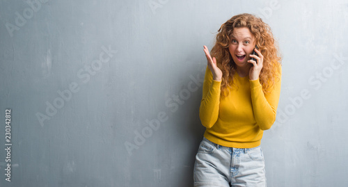 Young redhead woman over grey grunge wall talking on the phone very happy and excited, winner expression celebrating victory screaming with big smile and raised hands