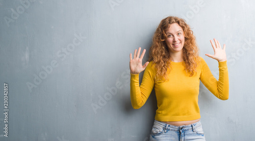 Young redhead woman over grey grunge wall showing and pointing up with fingers number ten while smiling confident and happy.