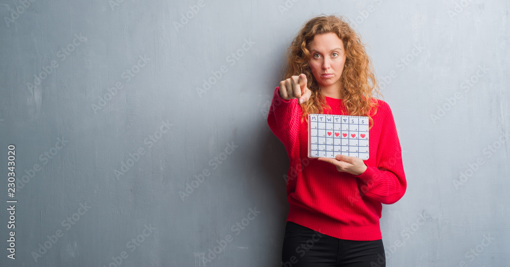 Young redhead woman over grey grunge wall holding period calendar pointing with finger to the camera and to you, hand sign, positive and confident gesture from the front Young redhead woman over grey grunge wall holding period calendar pointing with finger to the camera and to you, hand sign, positive and confident gesture from the front