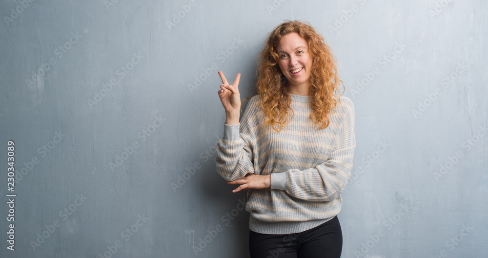 Young redhead woman over grey grunge wall smiling with happy face winking at the camera doing victory sign. Number two. Young redhead woman over grey grunge wall smiling with happy face winking at the camera doing victory sign. Number two.