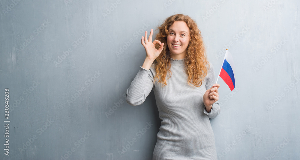 Young redhead woman over grey grunge wall holding flag of Russia doing ok sign with fingers, excellent symbol Young redhead woman over grey grunge wall holding flag of Russia doing ok sign with fingers, excellent symbol