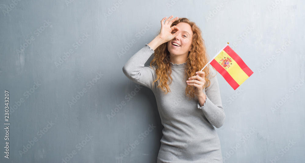 Young redhead woman over grey grunge wall holding flag of Spain with happy face smiling doing ok sign with hand on eye looking through fingers Young redhead woman over grey grunge wall holding flag of Spain with happy face smiling doing ok sign with hand on eye looking through fingers