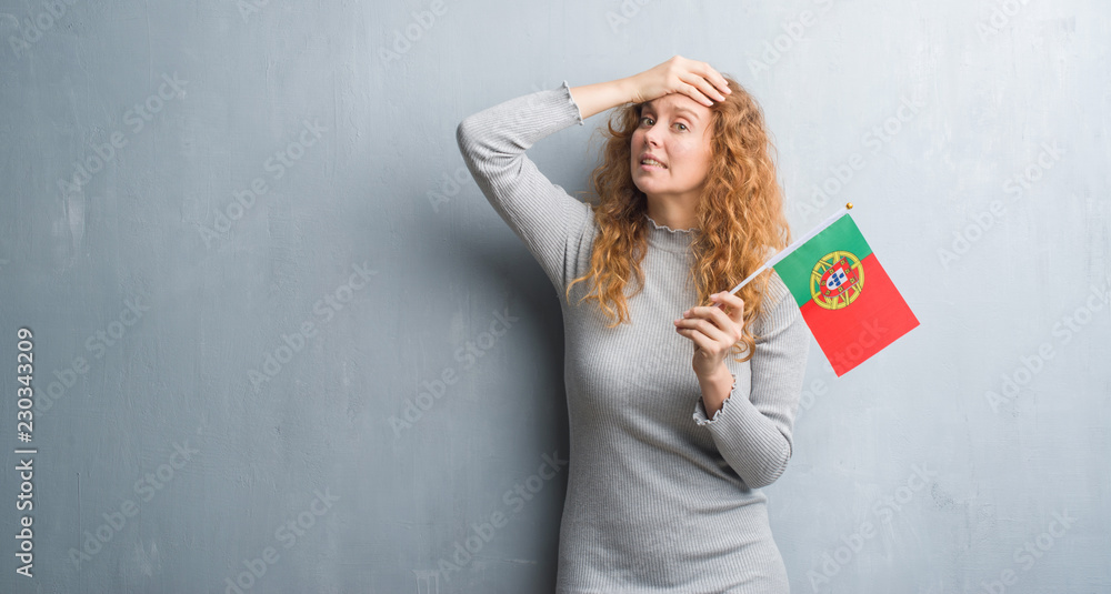 Young redhead woman over grey grunge wall holding flag of Portugal stressed with hand on head, shocked with shame and surprise face, angry and frustrated. Fear and upset for mistake. Young redhead woman over grey grunge wall holding flag of Portugal stressed with hand on head, shocked with shame and surprise face, angry and frustrated. Fear and upset for mistake.