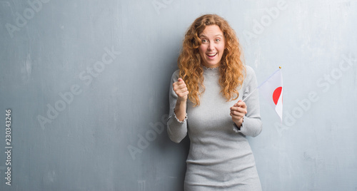 Young redhead woman over grey grunge wall holding flag of Japan screaming proud and celebrating victory and success very excited, cheering emotion