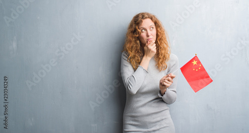 Young redhead woman over grey grunge wall holding flag of China serious face thinking about question, very confused idea