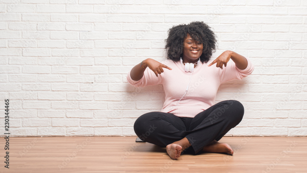 Young african american woman sitting on the floor wearing headphones looking confident with smile on face, pointing oneself with fingers proud and happy. Young african american woman sitting on the floor wearing headphones looking confident with smile on face, pointing oneself with fingers proud and happy.