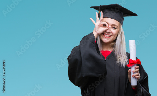 Young blonde woman wearing graduate uniform holding degree over isolated background with happy face smiling doing ok sign with hand on eye looking through fingers