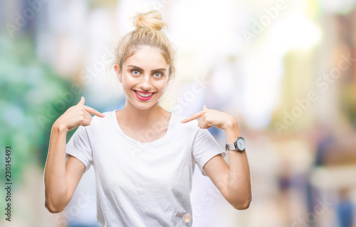 Young beautiful blonde woman wearing white t-shirt over isolated background looking confident with smile on face, pointing oneself with fingers proud and happy. © Krakenimages.com