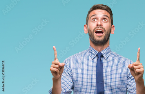 Young handsome business man over isolated background amazed and surprised looking up and pointing with fingers and raised arms.