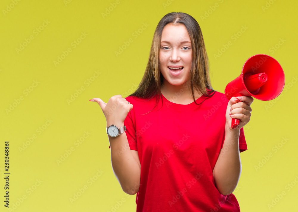 Young caucasian woman yelling through megaphone over isolated background pointing and showing with thumb up to the side with happy face smiling Young caucasian woman yelling through megaphone over isolated background pointing and showing with thumb up to the side with happy face smiling
