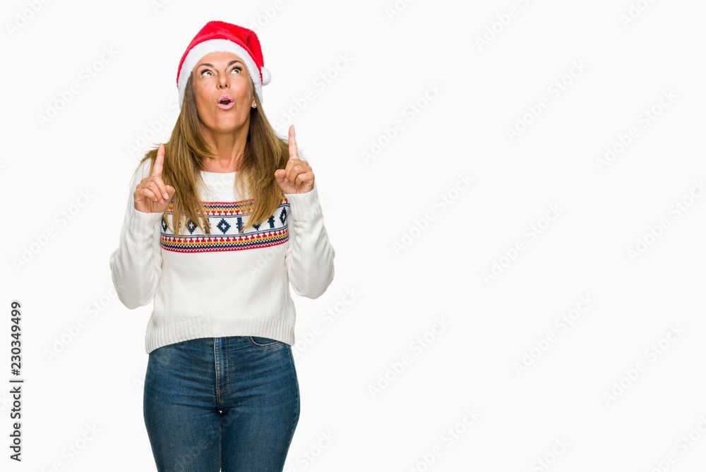Middle age adult woman wearing winter sweater and chrismat hat over isolated background amazed and surprised looking up and pointing with fingers and raised arms. Middle age adult woman wearing winter sweater and chrismat hat over isolated background amazed and surprised looking up and pointing with fingers and raised arms.