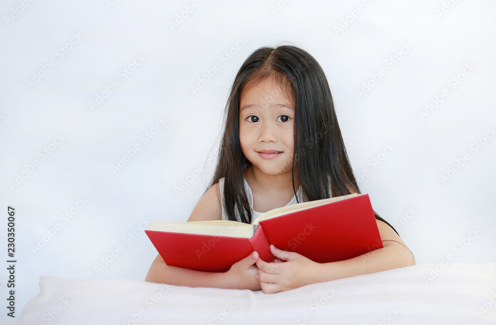 Smiling little Asian kid girl reading hardcover book lying with pillow on bed against white background. Smiling little Asian kid girl reading hardcover book lying with pillow on bed against white background.