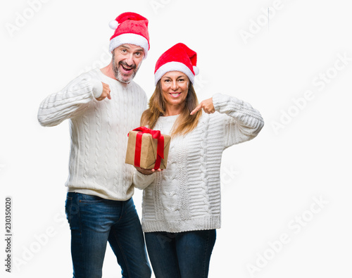 Middle age hispanic couple wearing christmas hat and holding gift over isolated background with surprise face pointing finger to himself