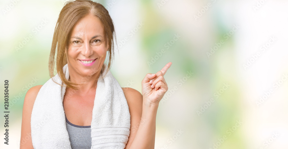 Beautiful middle age woman wearing sport clothes and a towel over isolated background with a big smile on face, pointing with hand and finger to the side looking at the camera.