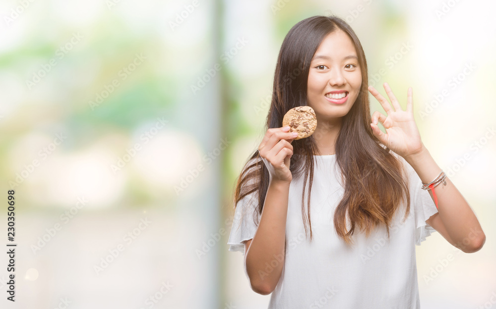 Young asian woman eating chocolate chip cookie over isolated background doing ok sign with fingers, excellent symbol Young asian woman eating chocolate chip cookie over isolated background doing ok sign with fingers, excellent symbol