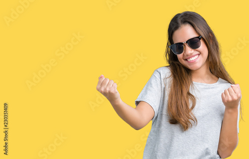 Young beautiful brunette woman wearing sunglasses over isolated background celebrating surprised and amazed for success with arms raised and open eyes. Winner concept.
