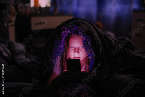 Little girl in bed under a blanket looking at the smartphone at night.