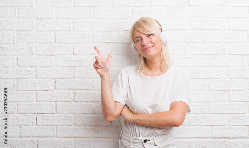 Adult caucasian woman over white brick wall wearing headphones with a big smile on face, pointing with hand and finger to the side looking at the camera. Adult caucasian woman over white brick wall wearing headphones with a big smile on face, pointing with hand and finger to the side looking at the camera.