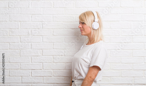Adult caucasian woman over white brick wall wearing headphones looking to side, relax profile pose with natural face with confident smile.