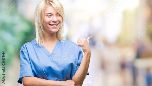 Young beautiful blonde nurse doctor woman over isolated background smiling with happy face looking and pointing to the side with thumb up. © Krakenimages.com