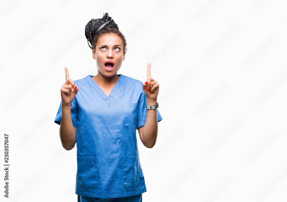 Young braided hair african american girl professional nurse over isolated background amazed and surprised looking up and pointing with fingers and raised arms. Young braided hair african american girl professional nurse over isolated background amazed and surprised looking up and pointing with fingers and raised arms.