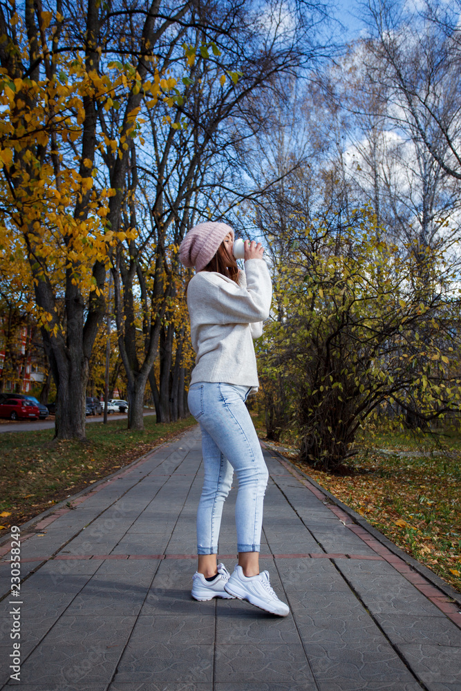 Fototapeta premium Happy young adult woman walking at beautiful autumn city street drinking coffee to go wearing warm coat and hat