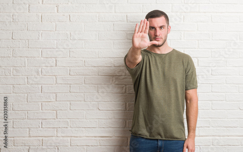 Young caucasian man standing over white brick wall doing stop sing with palm of the hand. Warning expression with negative and serious gesture on the face.