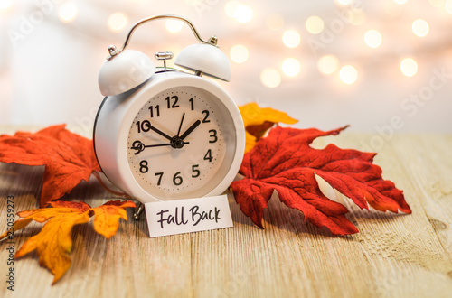 Fall Back Daylight Saving Time concept with white clock and autumn leaves, soft bokeh background on wooden board