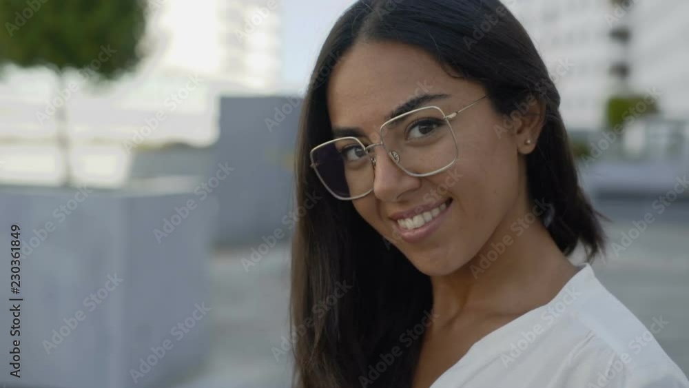 Beautiful Hispanic woman in eyeglasses looking at camera in city. Stylish smiling woman sitting on urban street. Positivity concept Beautiful Hispanic woman in eyeglasses looking at camera in city. Stylish smiling woman sitting on urban street. Positivity concept