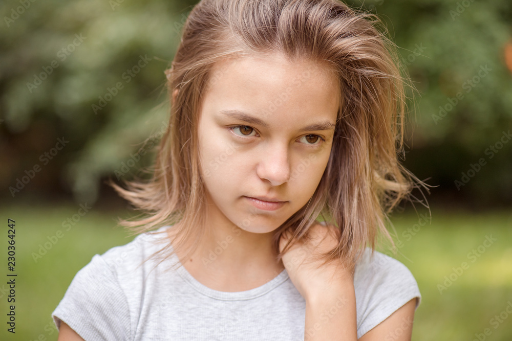 Fototapeta premium Emotional portrait of attractive caucasian little student girl with beautiful brown eyes in summer city park. Sad or modest child looking away - close-up, outdoors.