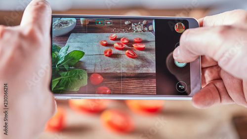 Sliced tomato on a wooden board. Woman's hands with a smartphone take off cooking salad. Food blogger concept