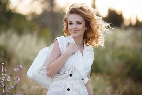Beautiful young woman with long curly hair, blonde, cute smile, dressed in a white dress with short sleeves, posing alone outdoors in the summer while strolling along a flowering green field.