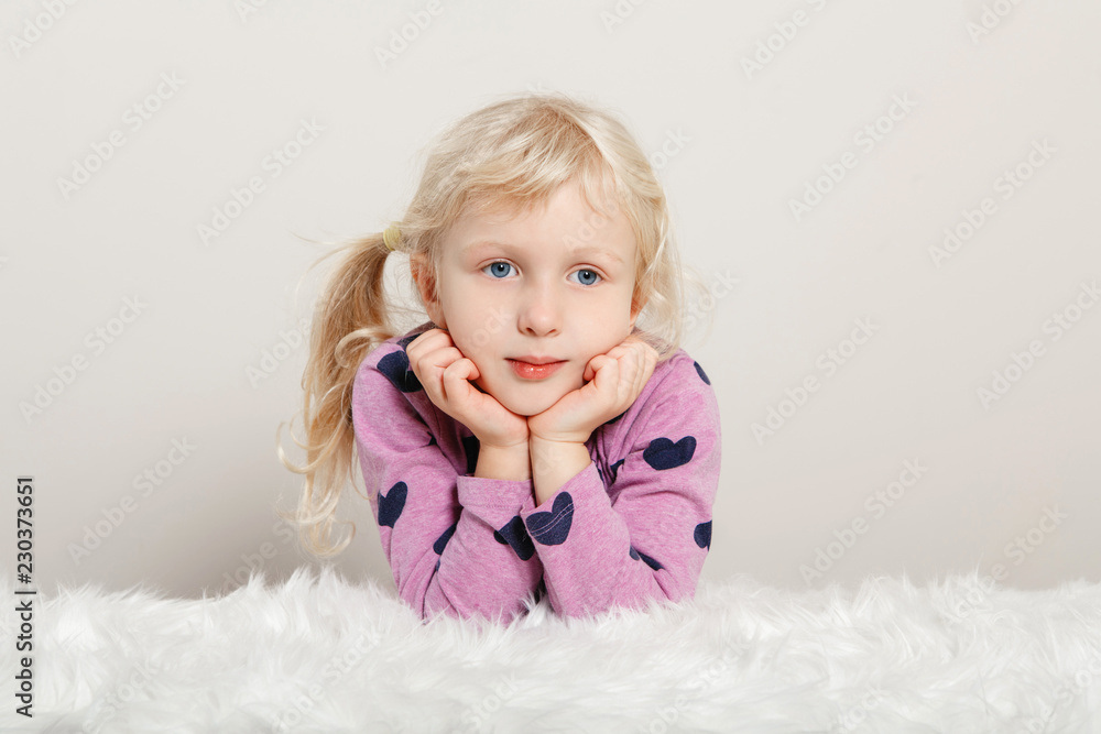 Closeup portrait of cute adorable white blonde Caucasian preschool girl in pink purple clothes sitting on white fur rug. Pensive child posing in studio on plain light background. Closeup portrait of cute adorable white blonde Caucasian preschool girl in pink purple clothes sitting on white fur rug. Pensive child posing in studio on plain light background.