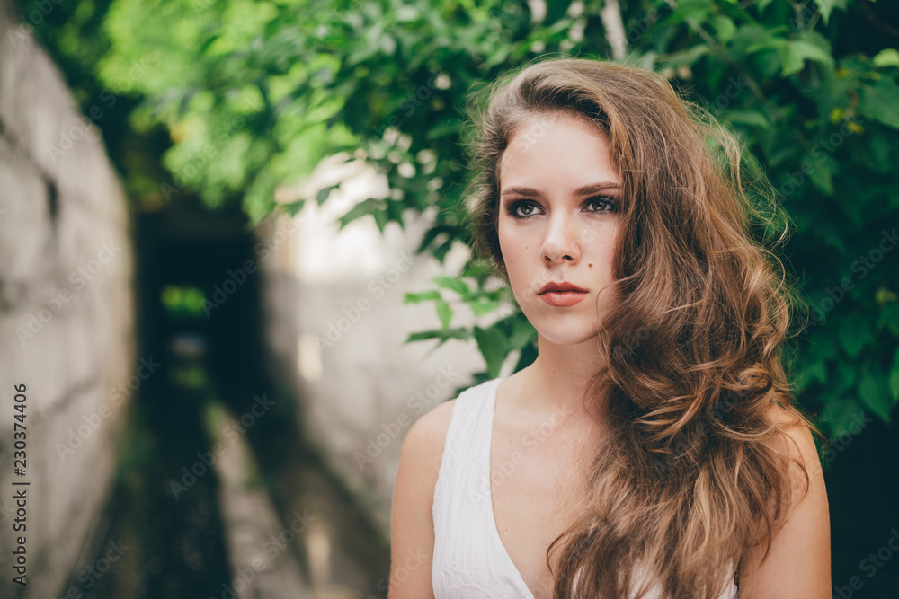 Beautiful moody girl with curly natural hair in white dress near green tree leaves. Summer beauty portrait. Sad lady in nature outdoors. Alone serious woman with pensive sight in long tunnel in forest Beautiful moody girl with curly natural hair in white dress near green tree leaves. Summer beauty portrait. Sad lady in nature outdoors. Alone serious woman with pensive sight in long tunnel in forest