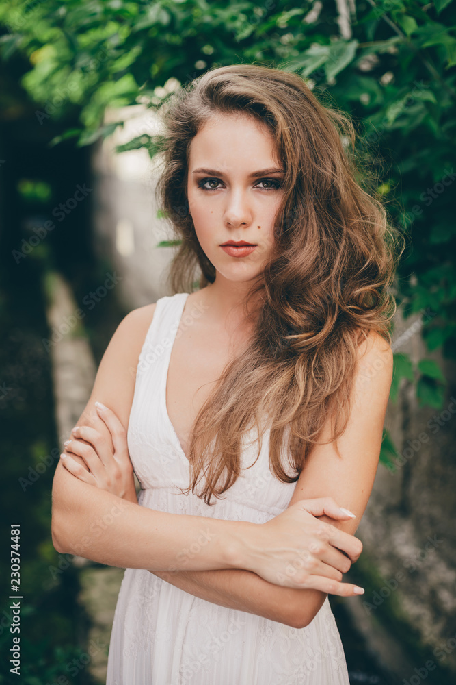 Beautiful moody girl with curly natural hair in white dress near green tree leaves. Summer beauty portrait. Sad lady in nature outdoors. Alone serious woman with pensive sight in long tunnel in forest Beautiful moody girl with curly natural hair in white dress near green tree leaves. Summer beauty portrait. Sad lady in nature outdoors. Alone serious woman with pensive sight in long tunnel in forest