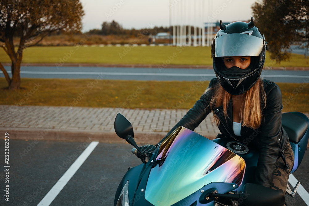 Blonde girl in protective helmet and black leather jacket sitting on motorbike, ready for race. People, extreme lifestyle, sports and adrenaline. Stylish confident female motor racer at parking lot