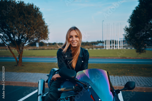 Outdoorimage of attractive young European woman with loose blonde hair sitting on blue motorbyke, dressed in camouflage jeans and black leather jacket, looking at camera with happy cute smile photo